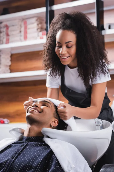 Lächelnder afrikanisch-amerikanischer Friseur hält Handtuch in der Nähe des Kopfes der Kundin im Salon — Stockfoto