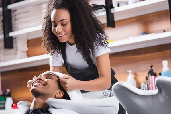 Coiffeur souriant tenant serviette près du client afro-américain et évier — Photo de stock