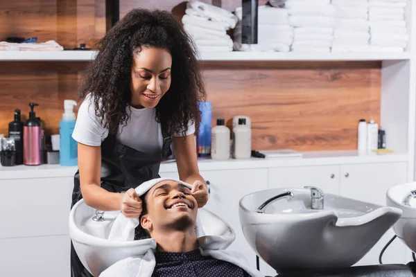 Joyeux homme afro-américain assis près de l'évier et coiffeur avec serviette dans le salon — Photo de stock