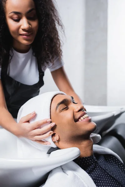 Jeune client afro-américain souriant près de l'évier et coiffeur avec serviette sur fond flou — Photo de stock