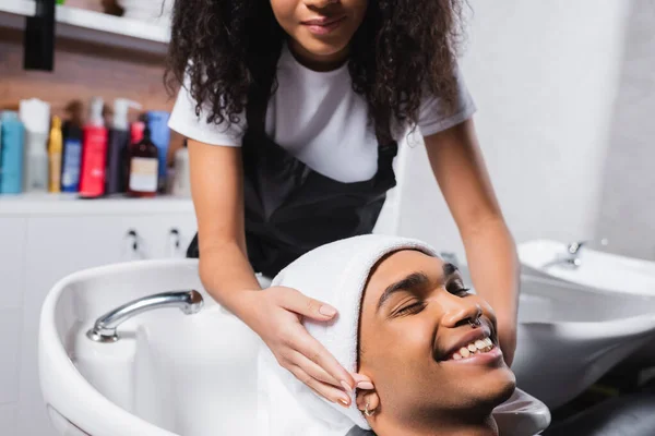Peluquería afroamericana envolviendo la cabeza del cliente sonriente en toalla - foto de stock