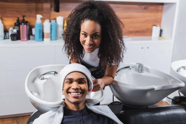 Alegre cabeleireiro afro-americano olhando para a câmera perto do cliente com toalhas — Fotografia de Stock