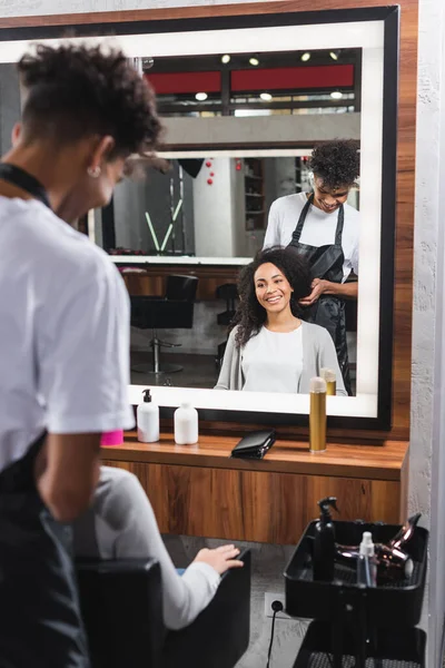 Mujer sonriente sentada cerca del peluquero afroamericano en primer plano borroso - foto de stock