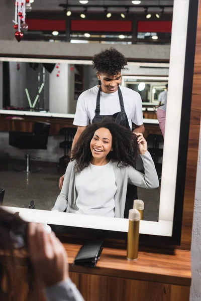 Sourire femme afro-américaine toucher les cheveux près de coiffeur dans le salon — Photo de stock