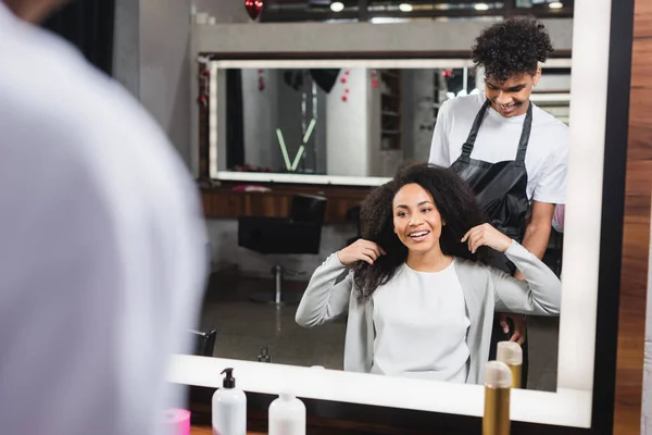 Fröhliche Friseurin in Schürze blickt auf Afroamerikanerin, die Haare berührt — Stockfoto