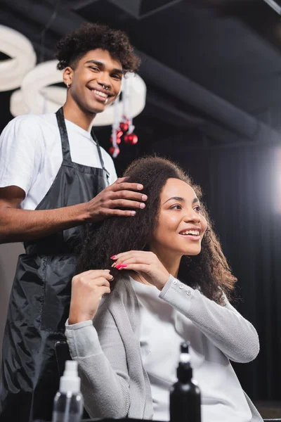 Low angle view of smiling african american woman sitting near blurred hairdresser — Stock Photo
