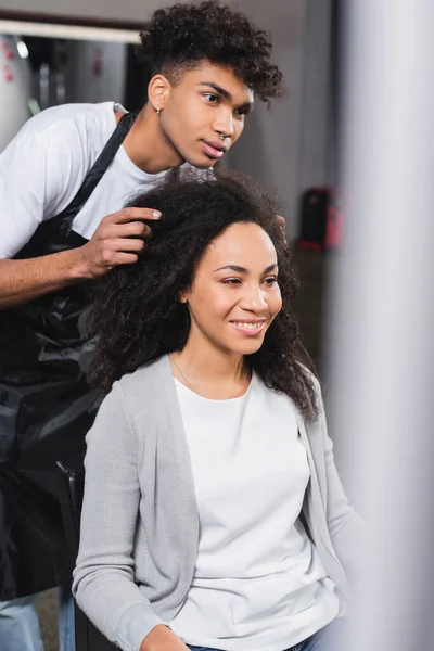 Mujer sonriente sentada cerca de peluquero afroamericano en delantal - foto de stock