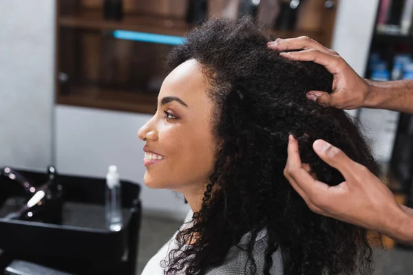 Vista laterale del cliente afroamericano sorridente seduto vicino parrucchiere toccare i capelli — Foto stock