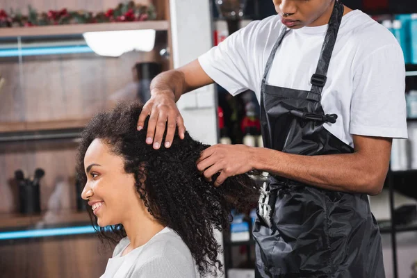 Smiling african american woman sitting near hairdresser in apron — Stock Photo