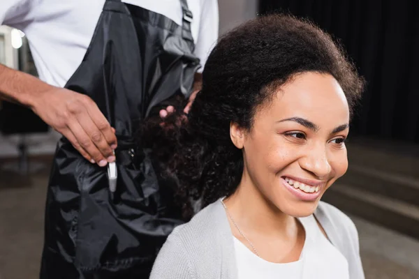 Alegre mujer afroamericana sentada cerca de peluquero con barreta sobre fondo borroso - foto de stock