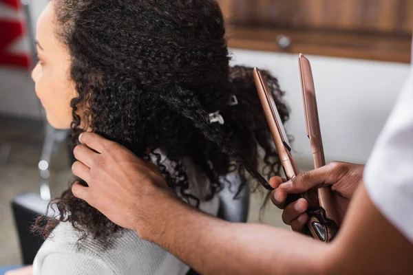 Peluquería afroamericana sosteniendo plancha de pelo cerca del cliente - foto de stock