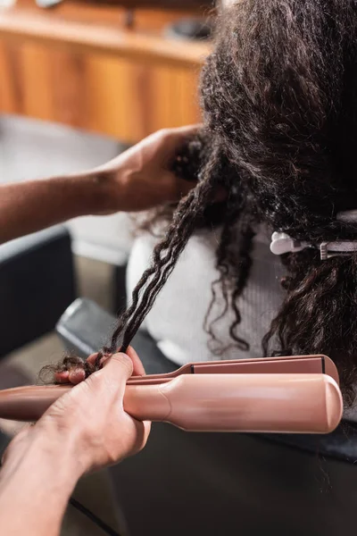 Vista recortada de peluquería afroamericana sosteniendo plancha de pelo cerca del cliente - foto de stock