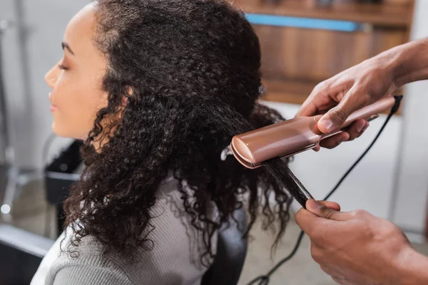 African american hairdresser using hair straightener on client — Stock Photo
