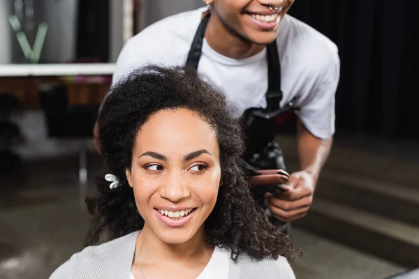Femme afro-américaine souriante regardant loin près de coiffeur flou avec fer à repasser — Photo de stock