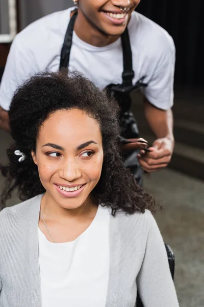 Sorridente donna africana americana guardando lontano vicino parrucchiere con piastra per capelli su sfondo sfocato — Foto stock