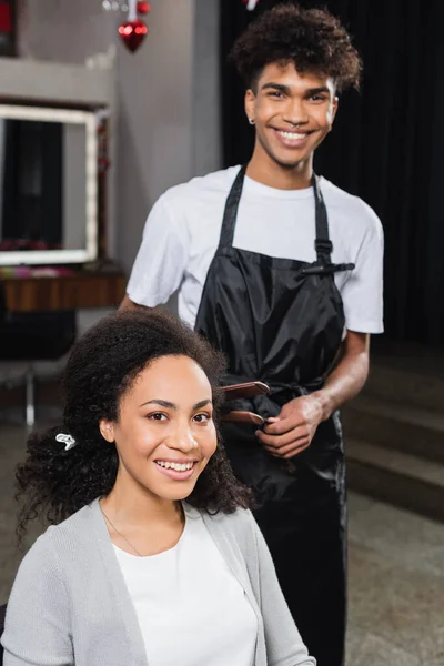 Cliente afroamericano positivo mirando la cámara cerca de estilista con plancha para el cabello - foto de stock