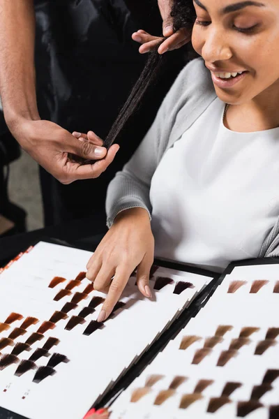 Улыбающийся африканский американский клиент указывает на образец цвета волос рядом с парикмахером — стоковое фото