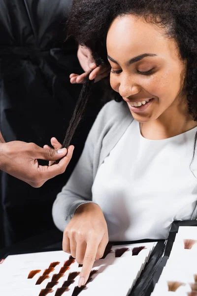 Alegre cliente afroamericano apuntando a la muestra de color de pelo cerca del estilista - foto de stock