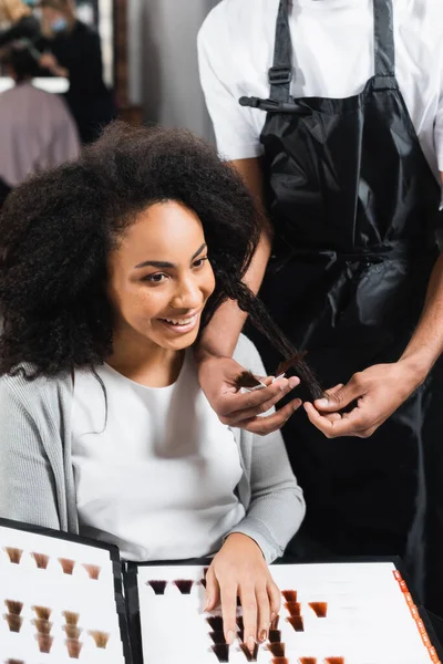 Sonriente mujer afroamericana sosteniendo la paleta de colores de cabello cerca del estilista - foto de stock