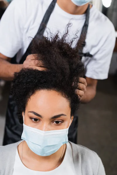 Mujer afroamericana en máscara médica sentada cerca de peluquero sobre fondo borroso - foto de stock