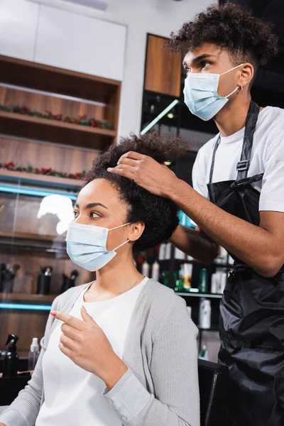 Coiffeur afro-américain en masque de protection touchant les cheveux de la jeune femme dans le salon — Photo de stock