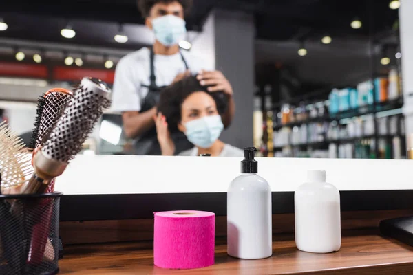 Spazzole per capelli, shampoo e carta collare vicino parrucchiere afroamericano sfocato che lavora con il cliente — Foto stock