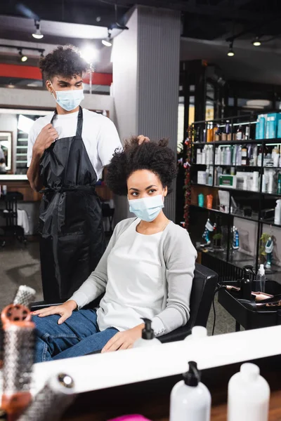 Peluquería afroamericana en máscara médica mirando al cliente cerca del espejo en el salón - foto de stock