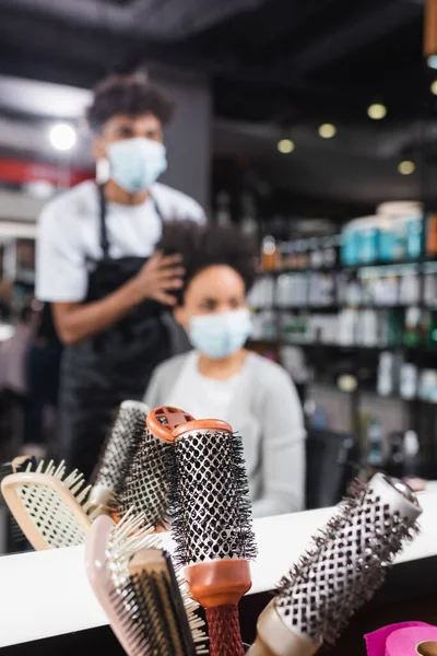 Escovas de cabelo perto de espelho e cliente afro-americano borrado em máscara médica e cabeleireiro — Fotografia de Stock