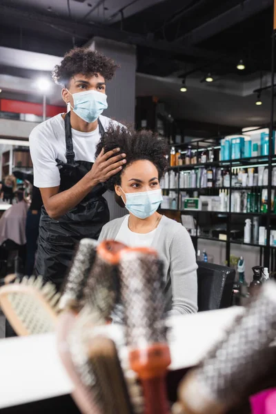 Coiffeur afro-américain en masque médical travaillant avec un client près de brosses floues — Photo de stock