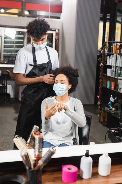 Parrucchiere afroamericano in maschera medica in piedi vicino al cliente utilizzando smartphone in salone — Foto stock