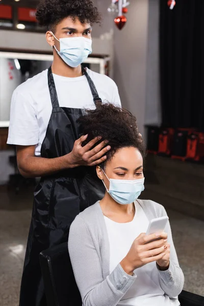 Mujer usando smartphone cerca de peluquero afroamericano en máscara médica - foto de stock