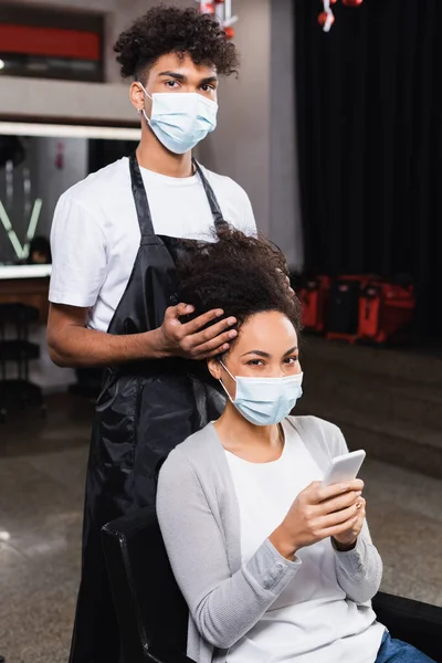 Mujer afroamericana con teléfono inteligente mirando a la cámara cerca de estilista en máscara médica - foto de stock