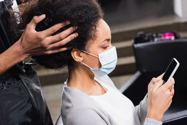 African american client in medical mask using smartphone near hairstylist in apron — Stock Photo