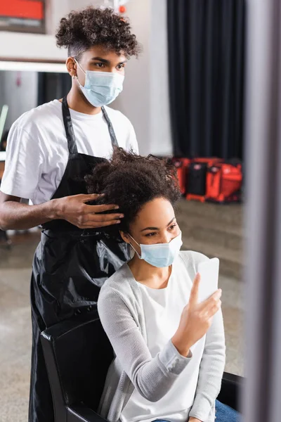 Cabeleireiro americano Africano tocando o cabelo do cliente com smartphone — Fotografia de Stock