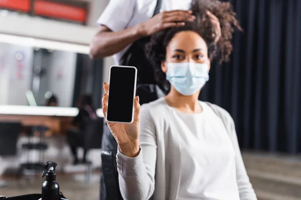 Smartphone con schermo bianco in mano del cliente afroamericano offuscato in maschera medica vicino al parrucchiere — Foto stock