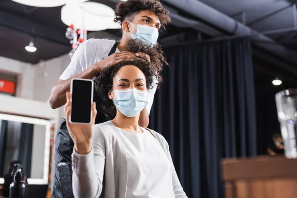 Смартфон с чистым экраном в руке африканской американки в медицинской маске рядом с парикмахером на размытом фоне — стоковое фото
