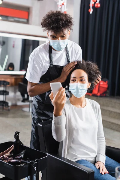 Cliente afroamericano en máscara protectora mostrando smartphone a peluquero - foto de stock