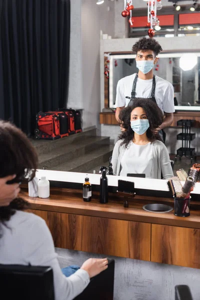 Peluquería afroamericana en máscara médica de pie cerca del cliente en el salón - foto de stock