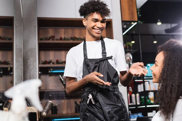 Cabeleireiro americano africano alegre apontando para o cliente em primeiro plano desfocado no salão — Fotografia de Stock