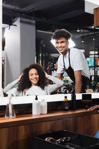 Smiling african american hairdresser and client looking at mirror in salon — Stock Photo