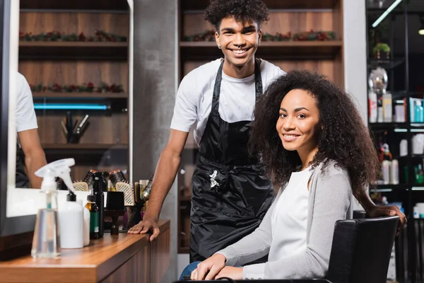 Cliente afroamericano e parrucchiere sorridente alla telecamera vicino allo specchio e alle attrezzature nel salone — Foto stock