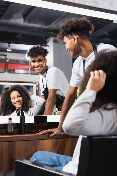 Sonriente peluquero afroamericano mirando el espejo cerca de la mujer tocando el pelo en primer plano borroso - foto de stock