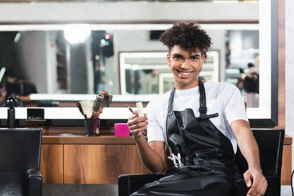 Smiling african american hairdresser holding comb and scissors in salon — Stock Photo
