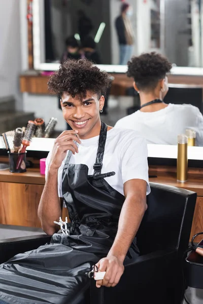 African american hairdresser with comb and scissors on blurred foreground smiling — Stock Photo