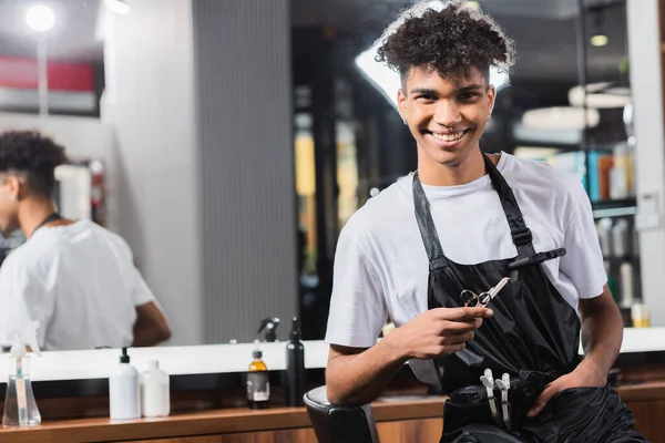 Молодой африканский американский парикмахер держит ножницы и расческу возле стула — стоковое фото