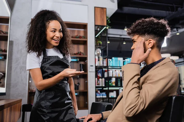 Sonriente peluquero afroamericano en delantal apuntando al cliente - foto de stock