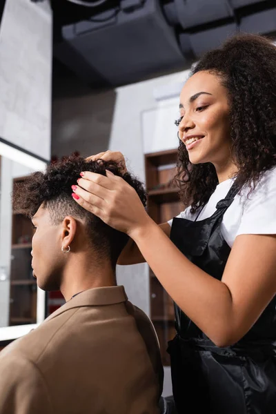 Smiling african american hairdresser looking at hair of man in salon — Stock Photo