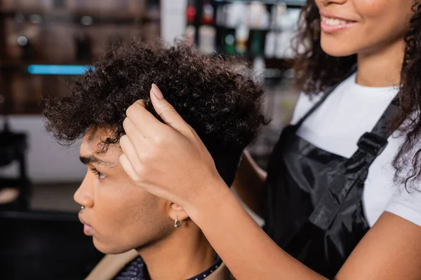 Улыбающийся парикмахер трогает кудрявые волосы молодого африканского американца в салоне — стоковое фото