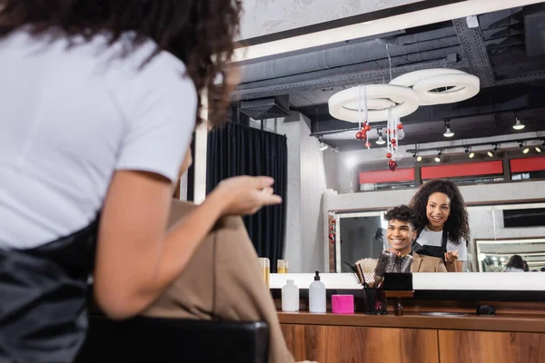 Peluquería afroamericana sonriente señalando con la mano cerca del cliente y espejo en el salón - foto de stock