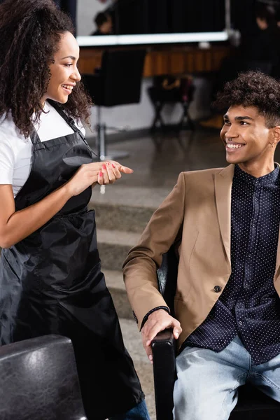 Happy african american hairdresser looking at man in chair — Stock Photo